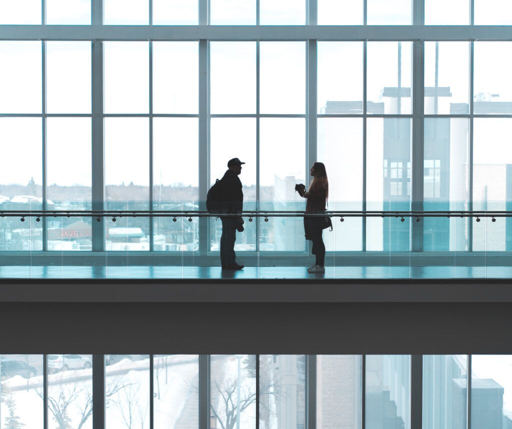 Two people standing in front of large windows in an office building, enjoying the view and discussing work.
