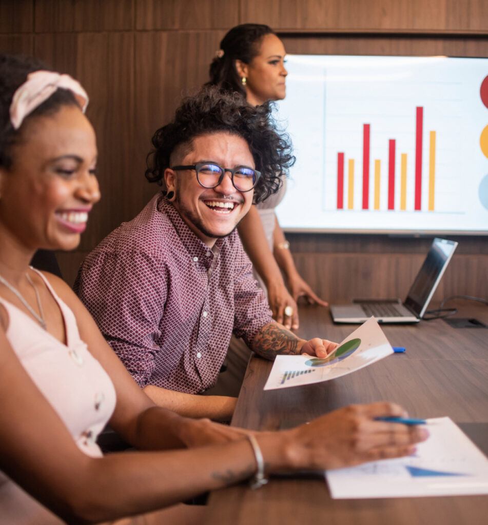 Three people sitting at a table, looking at a chart on the wall, with big smiles during a meeting.