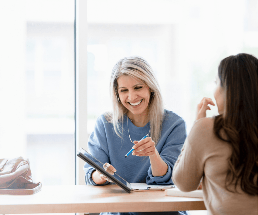 Two women engaged in a meeting, sitting at a table with a tablet and pen, discussing important matters.